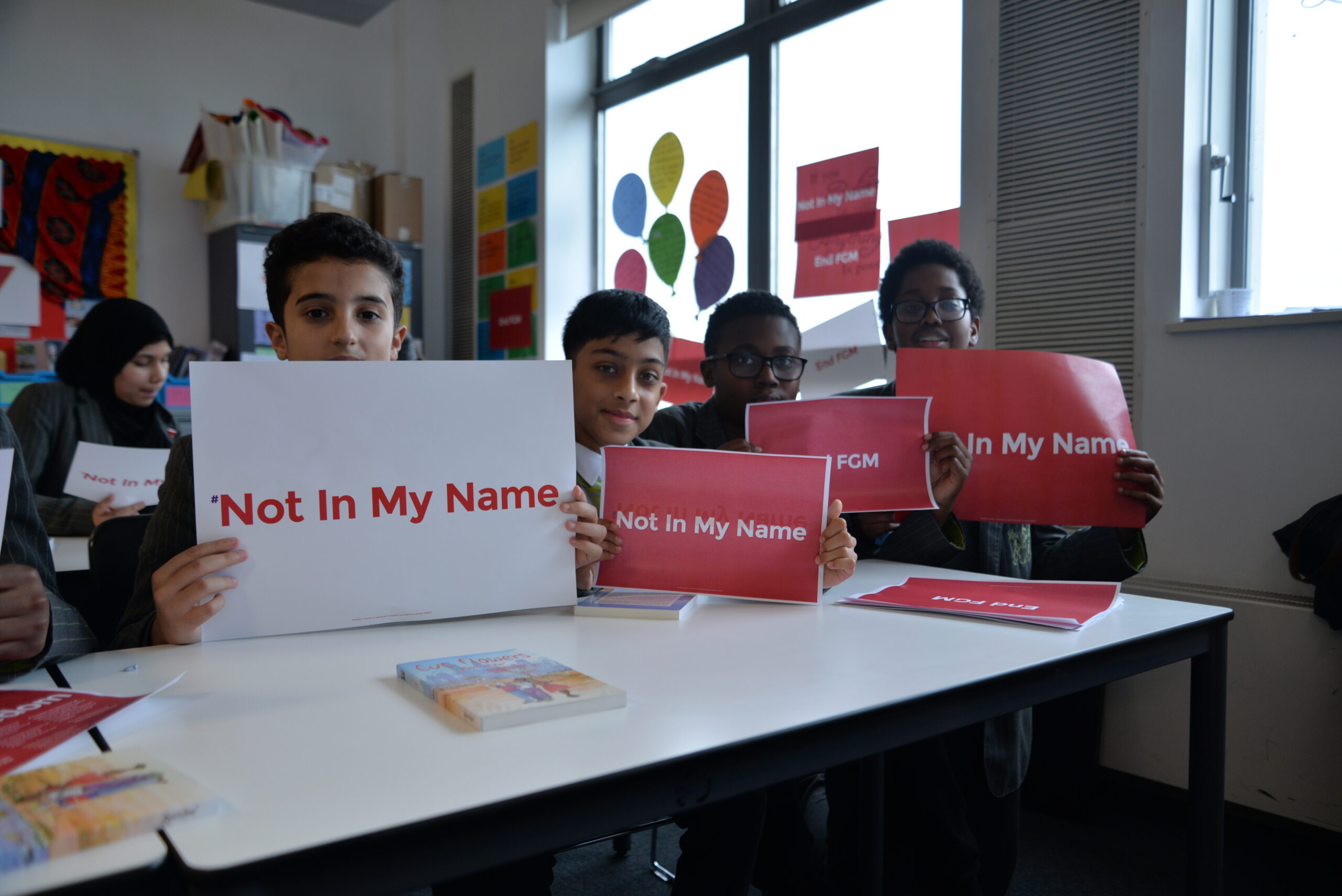 Young people in a classroom holding Not in My Name and End FGM signs